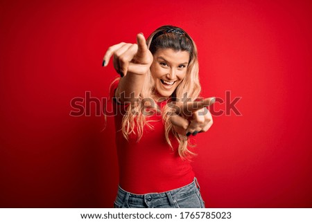 Young beautiful blonde woman wearing casual t-shirt standing over isolated red background pointing to you and the camera with fingers, smiling positive and cheerful