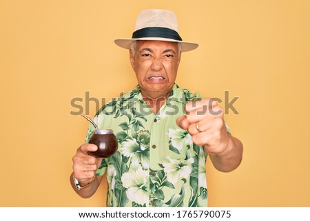 Middle age senior grey-haired man wearing summer hat drinking traditional mate drink annoyed and frustrated shouting with anger, crazy and yelling with raised hand, anger concept