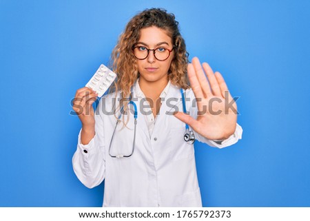 Young beautiful blonde doctor woman with blue eyes wearing stethoscope holding pills with open hand doing stop sign with serious and confident expression, defense gesture