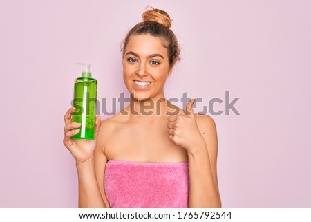 Beautiful blonde woman with blue eyes wearing towel shower after bath holding aloe vera gel happy with big smile doing ok sign, thumb up with fingers, excellent sign