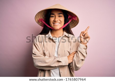 Young beautiful woman wearing traditional conical asian hat over isolated pink background with a big smile on face, pointing with hand and finger to the side looking at the camera.