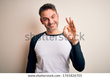 Young handsome man wearing casual t-shirt standing over isolated white background smiling positive doing ok sign with hand and fingers. Successful expression.