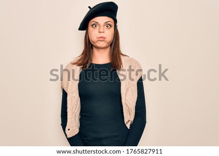 Young beautiful brunette woman wearing casual french beret over white background puffing cheeks with funny face. Mouth inflated with air, crazy expression.