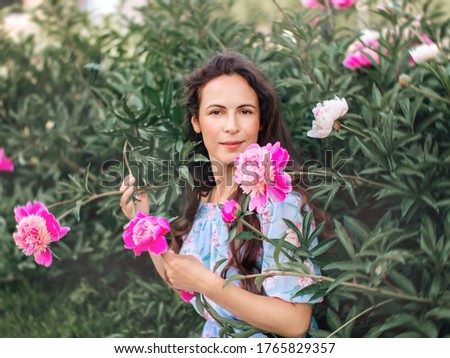 A brunette girl in a blue dress holds pink peony flowers in her hands, sitting on the grass in the Park near the flowers