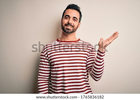 Young handsome man with beard wearing casual striped t-shirt standing over white background smiling cheerful presenting and pointing with palm of hand looking at the camera.