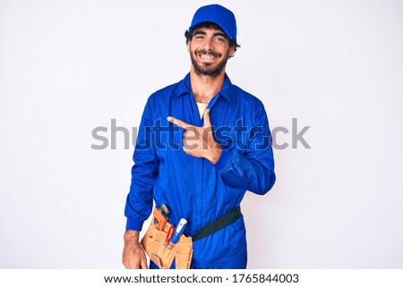 Handsome young man with curly hair and bear weaing handyman uniform cheerful with a smile on face pointing with hand and finger up to the side with happy and natural expression 
