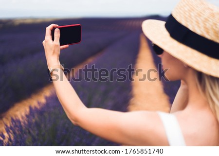 Side view of crop young female in hat making selfie while standing during trip on lavender meadows in Provence in France
