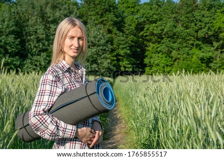 young blonde woman in a shirt holds a sports Mat in the middle of a field of green rye on a Sunny summer day