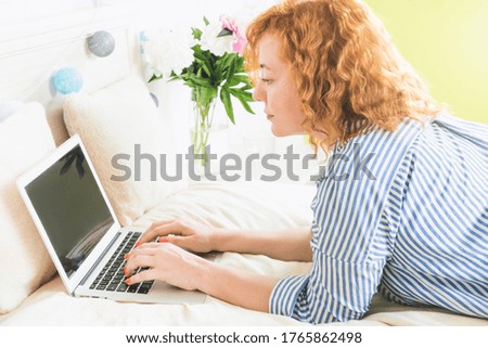 A woman with wavy red hair lies on the bed and works on a laptop. Work from home, distance education.