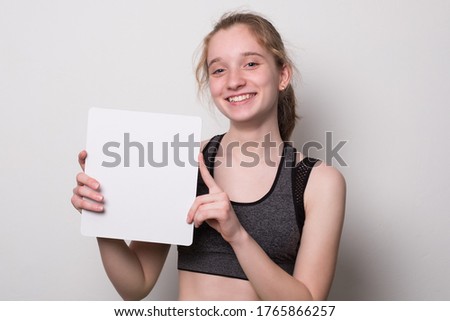 Positive girl holds a blank poster for text. Teen girl with a smile holds a white sheet for text.