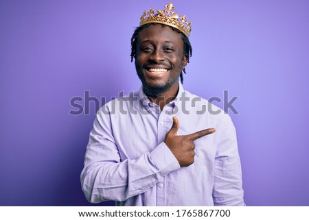 Young african american man wearing golden crown of king over isolated purple background cheerful with a smile of face pointing with hand and finger up to the side with happy and natural expression
