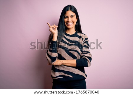 Young beautiful hispanic woman wearing animal print sweater over pink background with a big smile on face, pointing with hand and finger to the side looking at the camera.