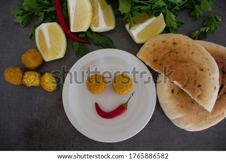 Smiling face made of food. Plate of falafels, pita bread, lemon and pepper on dark grey table. Traditional Israeli meal top view. 