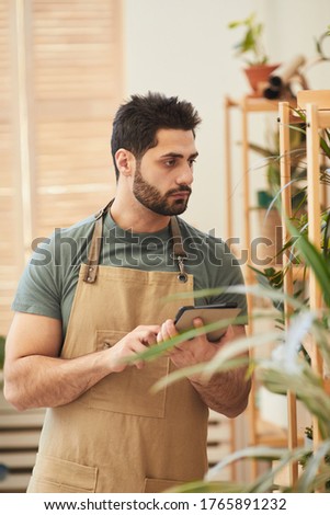 Vertical waist up portrait of handsome bearded man using digital tablet while counting stock in flower store