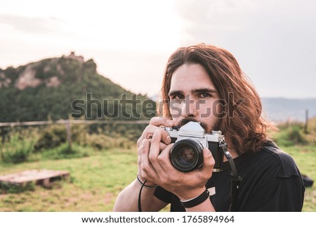 Adventurous young man looking at camera. He's looking over his camera. Behind him is a mountain with a building on top.