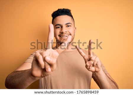 Young handsome man wearing casual t-shirt standing over isolated yellow background smiling looking to the camera showing fingers doing victory sign. Number two.
