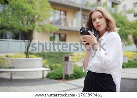 Photographer girl walks around the city with a reflex camera. A young blonde learns to take pictures. The model is looking at the camera and holding a camera in her hands. Photo Freelance Courses.