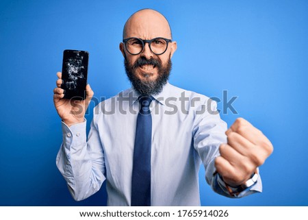 Handsome bald business man with beard holding broken smartphone showing cracked screen annoyed and frustrated shouting with anger, crazy and yelling with raised hand, anger concept