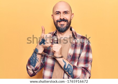 Young handsome man wearing prisoner handcuffs smiling happy and positive, thumb up doing excellent and approval sign 