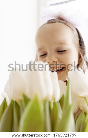 Close up charming little girl smiling, holding white tulips near the window, day light reflecting on her beautiful face