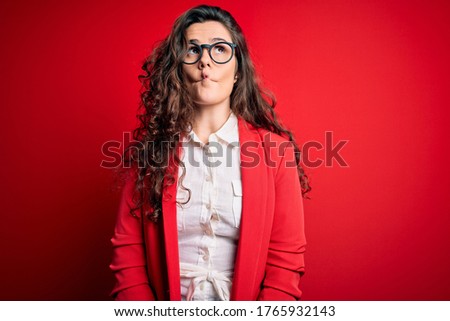 Young beautiful woman with curly hair wearing jacket and glasses over red background making fish face with lips, crazy and comical gesture. Funny expression.
