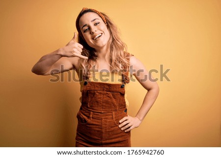 Young beautiful blonde woman wearing overalls and diadem standing over yellow background smiling doing phone gesture with hand and fingers like talking on the telephone. Communicating concepts.