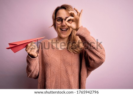 Young beautiful blonde woman wearing glasses holding paper airplane over pink background with happy face smiling doing ok sign with hand on eye looking through fingers