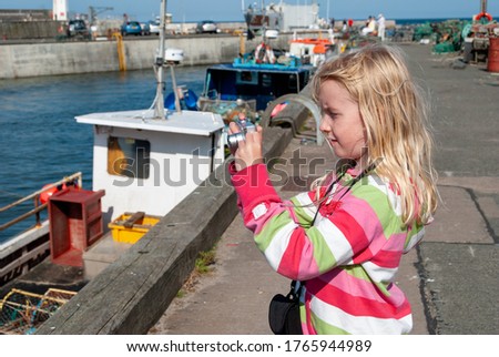 Close up of a young blonde girl taking a photo of an English harbour.