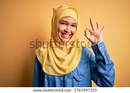 Young beautiful woman with curly hair wearing arab traditional hijab over yellow background smiling positive doing ok sign with hand and fingers. Successful expression.