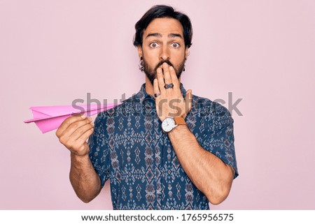 Young handsome hispanic bohemian hipster man with bear holding paper plane cover mouth with hand shocked with shame for mistake, expression of fear, scared in silence, secret concept