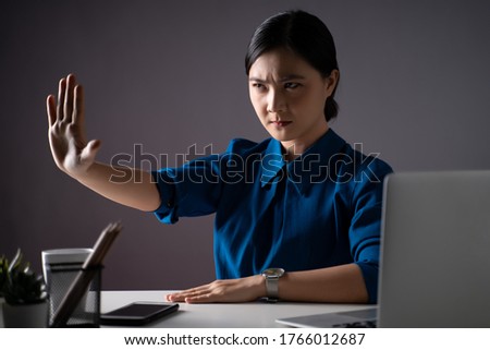 Asian woman in blue shirt working on a laptop at office, showing hand making stop sign, isolated on background. Low key.
