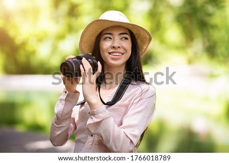 Outdoor Hobbies. Happy asian woman traveler in wicker hat taking photo with modern camera, enjoying landscapes
