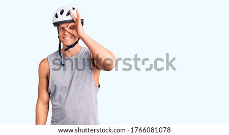 Young handsome man wearing bike helmet doing ok gesture with hand smiling, eye looking through fingers with happy face. 