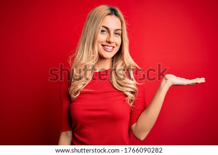 Young beautiful blonde woman wearing casual t-shirt standing over isolated red background smiling cheerful presenting and pointing with palm of hand looking at the camera.