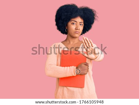 Young african american girl holding book with open hand doing stop sign with serious and confident expression, defense gesture 