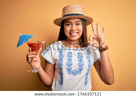 Young asian girl on vacation wearing summer hat drinking cocktail over yellow background doing ok sign with fingers, excellent symbol
