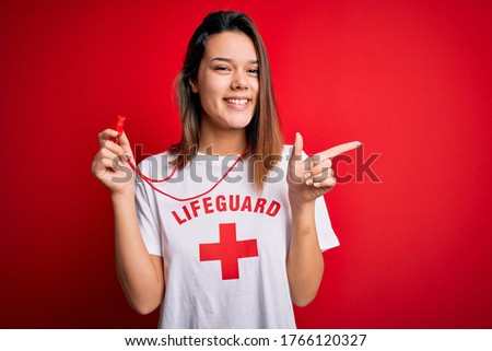 Young beautiful brunette lifeguard girl wearing t-shirt with red cross using whistle cheerful with a smile on face pointing with hand and finger up to the side with happy and natural expression