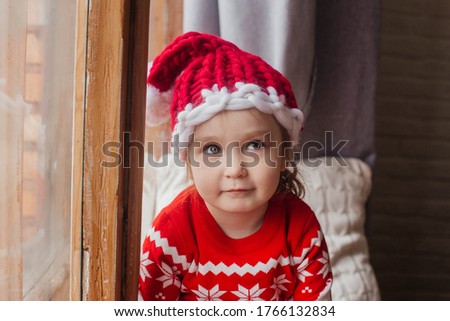 Little cute girl in a New Year hat and a sweater sits at the window.
Merry christmas and happy new year concept