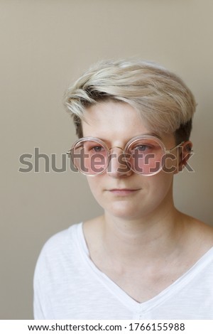 Close-up portrait of young trendy woman with short blonde haircut wearing pink sunglasses over neutral beige background, looking away 