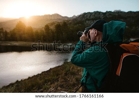 Old man on hiking vacation taking photographs of beautiful view with a digital camera. senior man standing by the river in forest and taking photos.