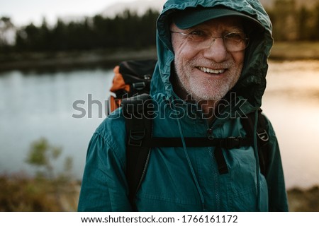 Portrait of a senior man carrying a backpack looking at camera and smiling. Fit old man on a hiking trip with river in background. Royalty-Free Stock Photo #1766161172