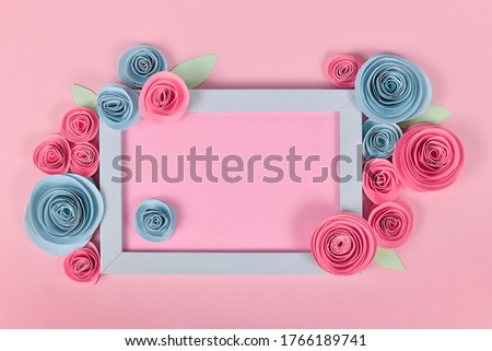 Blue and pink flower flat lay with empty picture frame surrounded by romantic paper craft roses on pastel pink background