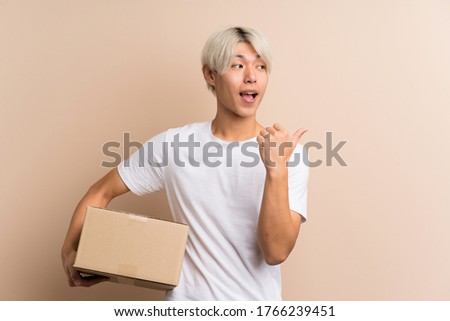 Young asian man over isolated background holding a box to move it to another site and pointing side