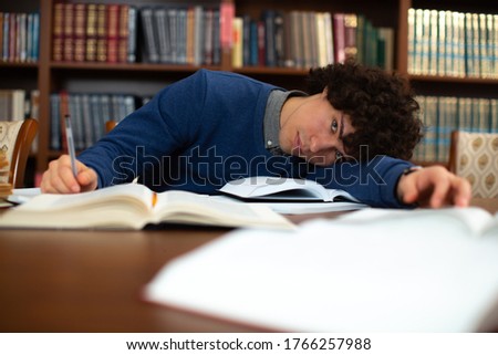 A tired curly-haired student in a grey-collared blue sweater, lay on his arm tired of homework and looking at the camera in the library