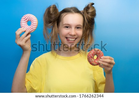 Charming girl on a blue background holds delicious donuts in her hands. Photo in pastel colors. Funny hairstyle and facial expressions. Tongue. Portrait. Horizontal photo