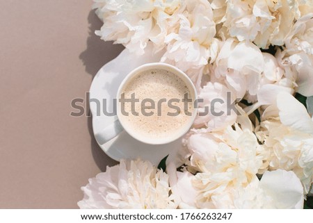 Coffee in a white mug surrounded by lush peonies on a light beige background. With place for text. Coffee and flowers. 