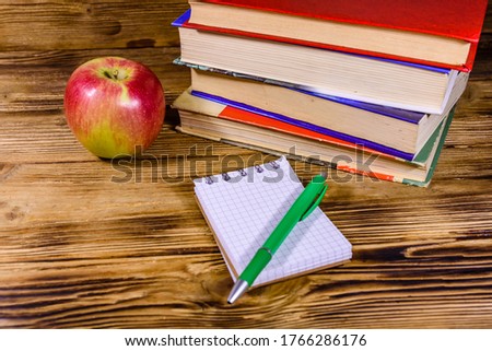Ripe apple, opened small notepad and ball pen on a wooden table in front of stack of books
