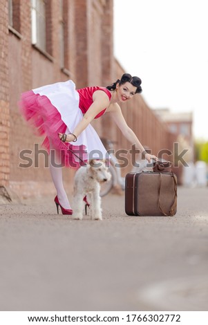 A girl in a dress and hairstyle in the style of the 40-50s on the city street with a dog breed Fox Terrier on a leash on a sunny day. Retro style photo. 