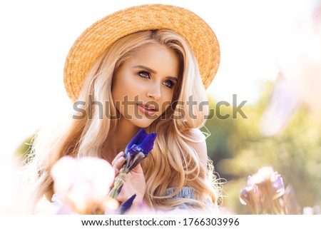 Beautiful blonde woman with wavy long hair wearing summer hat and posing near the flowers
