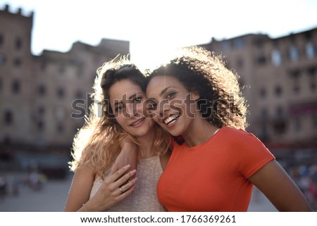 A blonde and black-haired lady hugging each other behind the buildings showing happiness and excitement